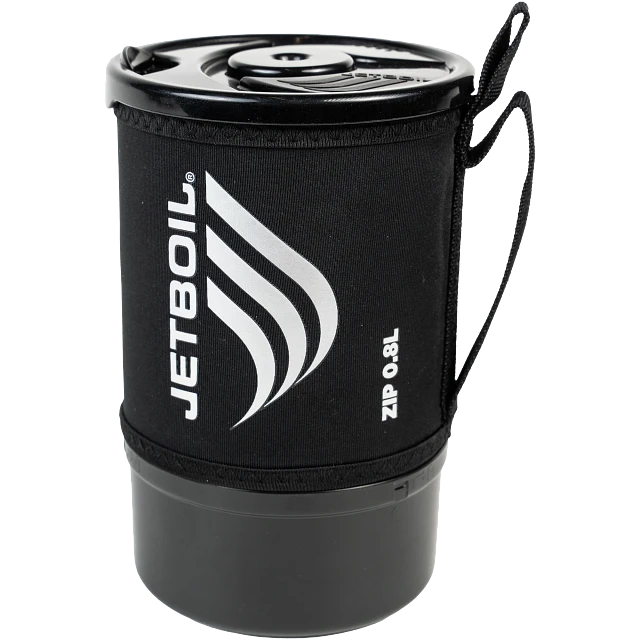 A Jetboil Zip 0.8L cooking system set up on a rocky surface outdoors, heating water with visible steam rising from the lid. A camper stands nearby, preparing a meal.