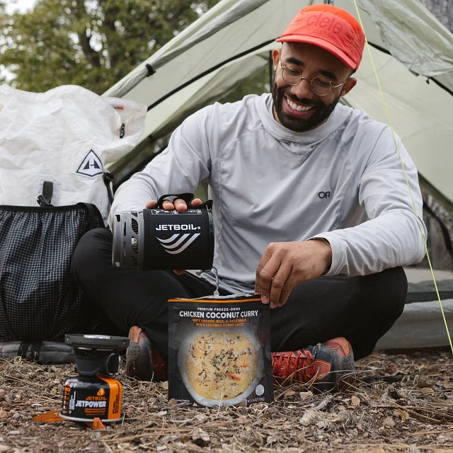 A camper using the Jetboil Zip 0.8L to prepare a hot drink at a campsite with mountains in the background. The compact system is placed on a flat surface, with a backpack and tent visible nearby.