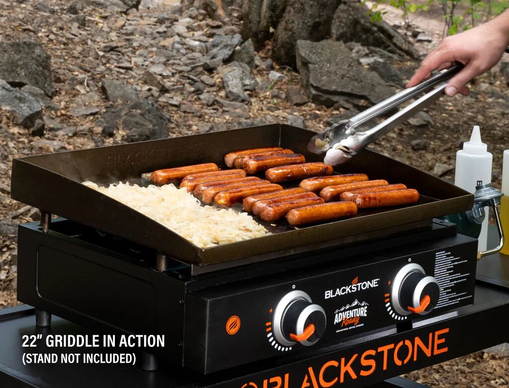 Blackstone griddle with hot dogs and tongs on a rocky outdoor surface