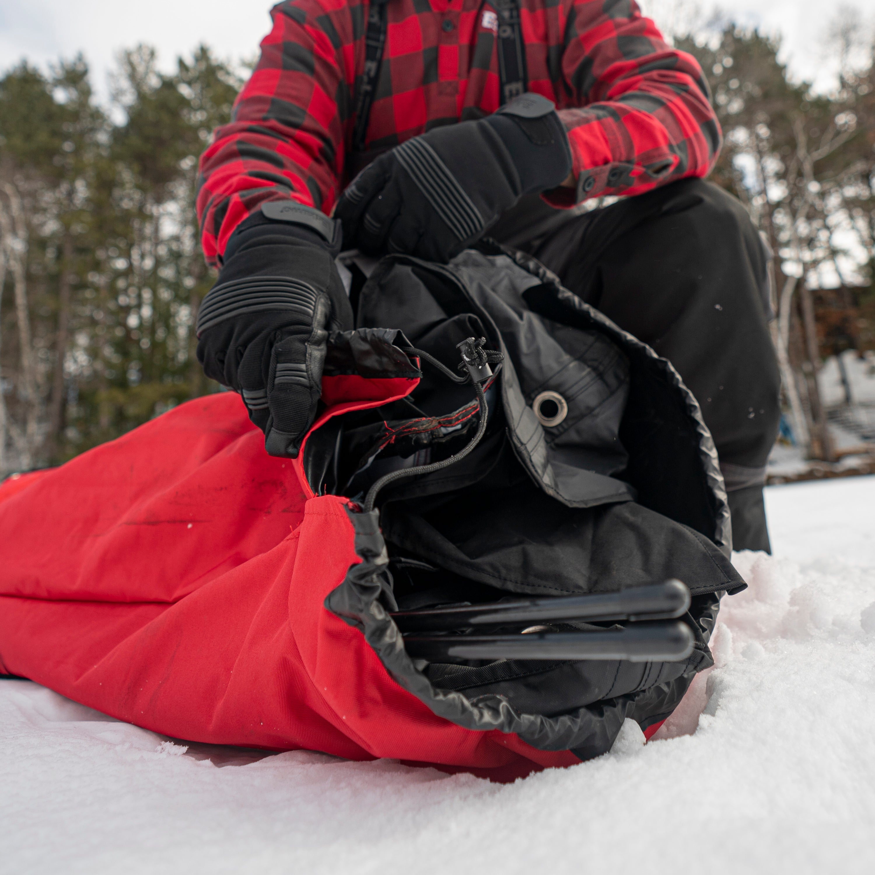 Person in red plaid jacket and black pants sitting in snow with a red sleeping bag and black backpack.