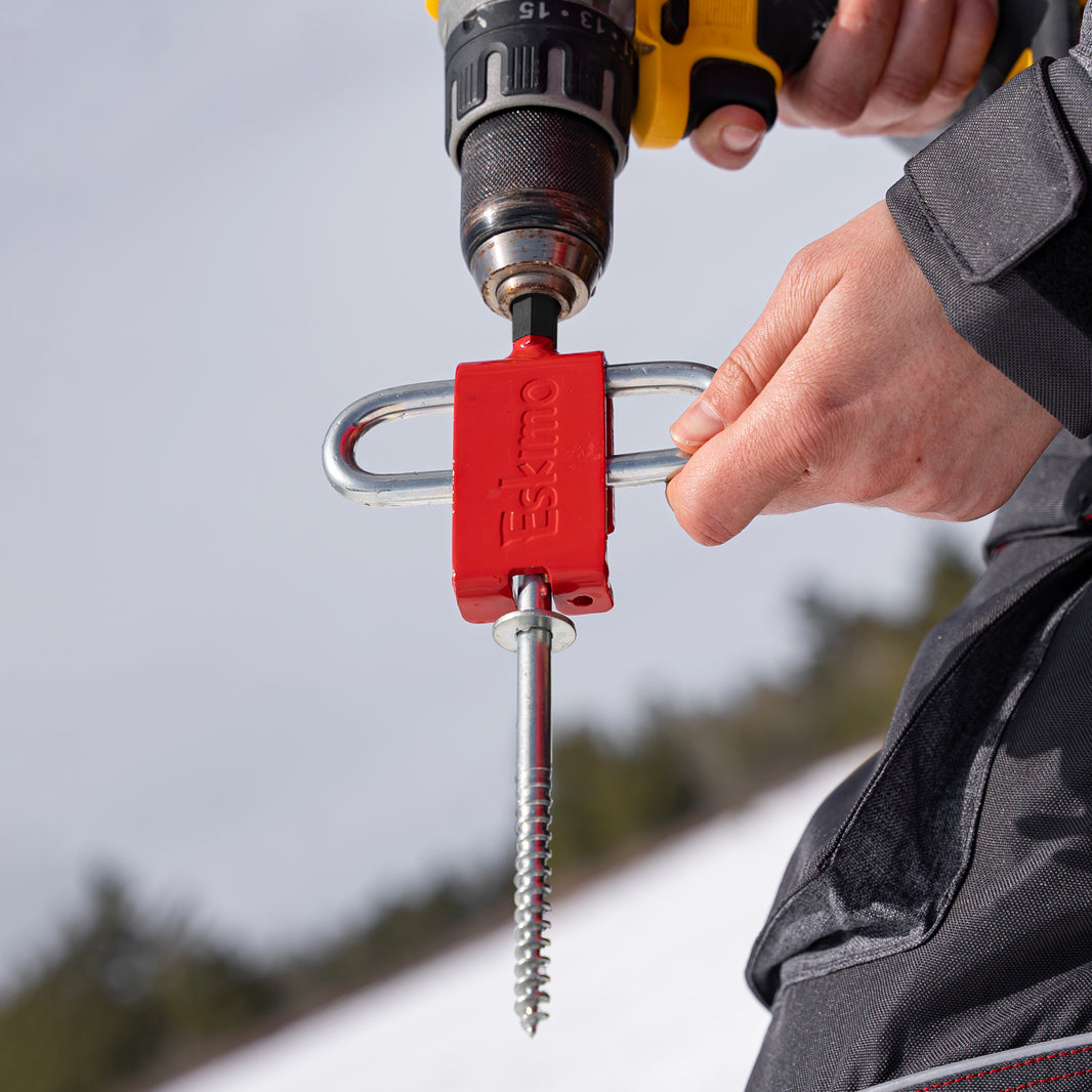 Person using a drill with a red ski anchor attachment against a blurred outdoor background