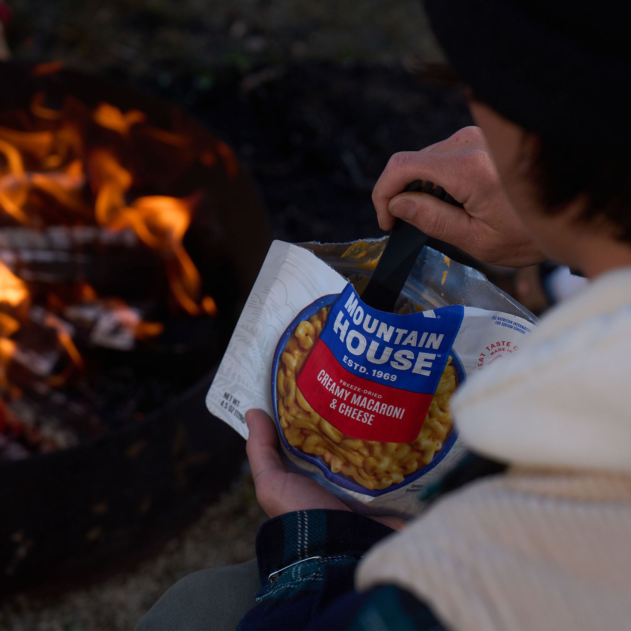 Person holding a Mountain House package of creamy macaroni and cheese in front of a campfire.