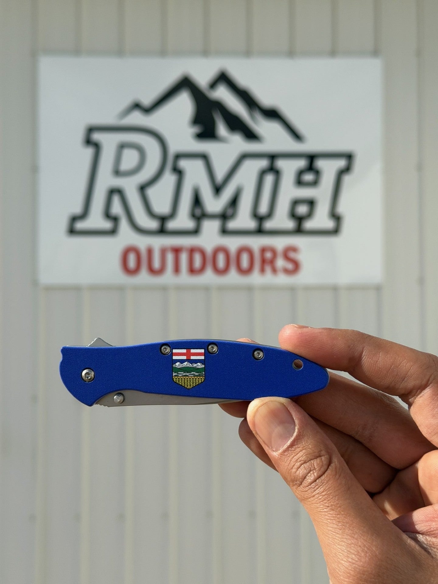 Blue folding knife held by a hand with RMH Outdoors logo in the background