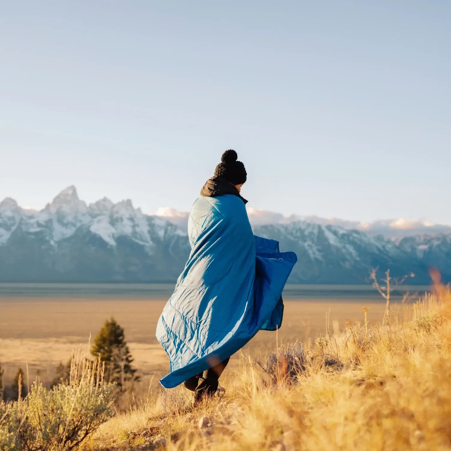Person wrapped in a blue blanket standing in a field with mountains in the background