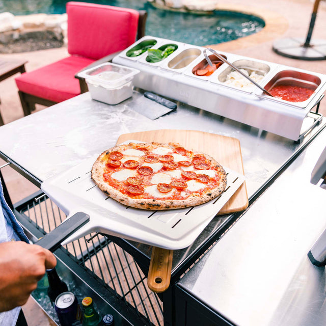 Personal pizza on a wooden board with pizza-making supplies on a table outdoors.