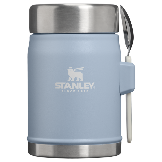 A light blue Stanley insulated container with a stainless steel lid and base, featuring a white Stanley logo on the front and a white-handled spoon attached on the side, shown against a plain white background.