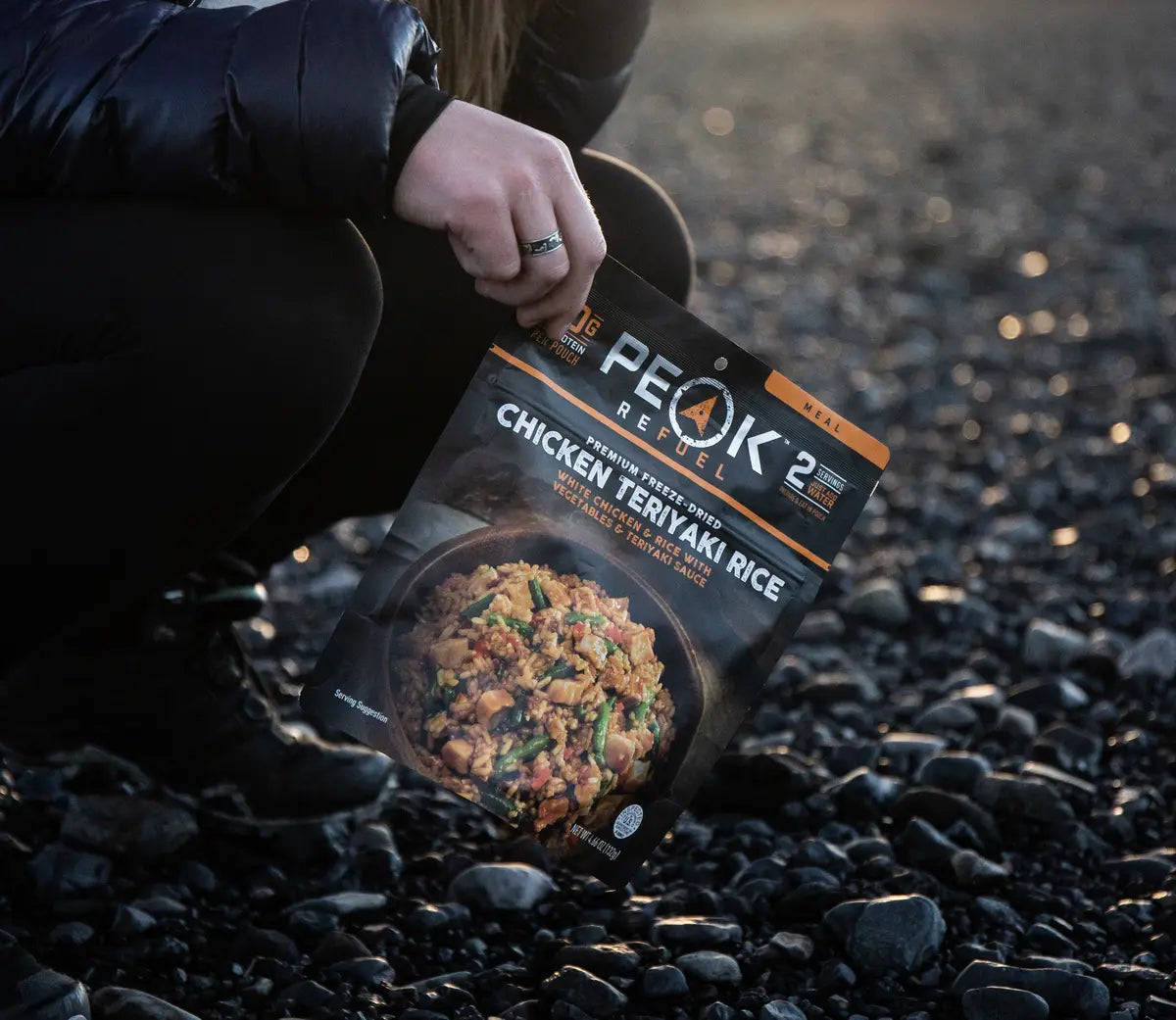 Person holding a package of Peak Rebel Chicken Teriyaki Rice on a rocky surface
