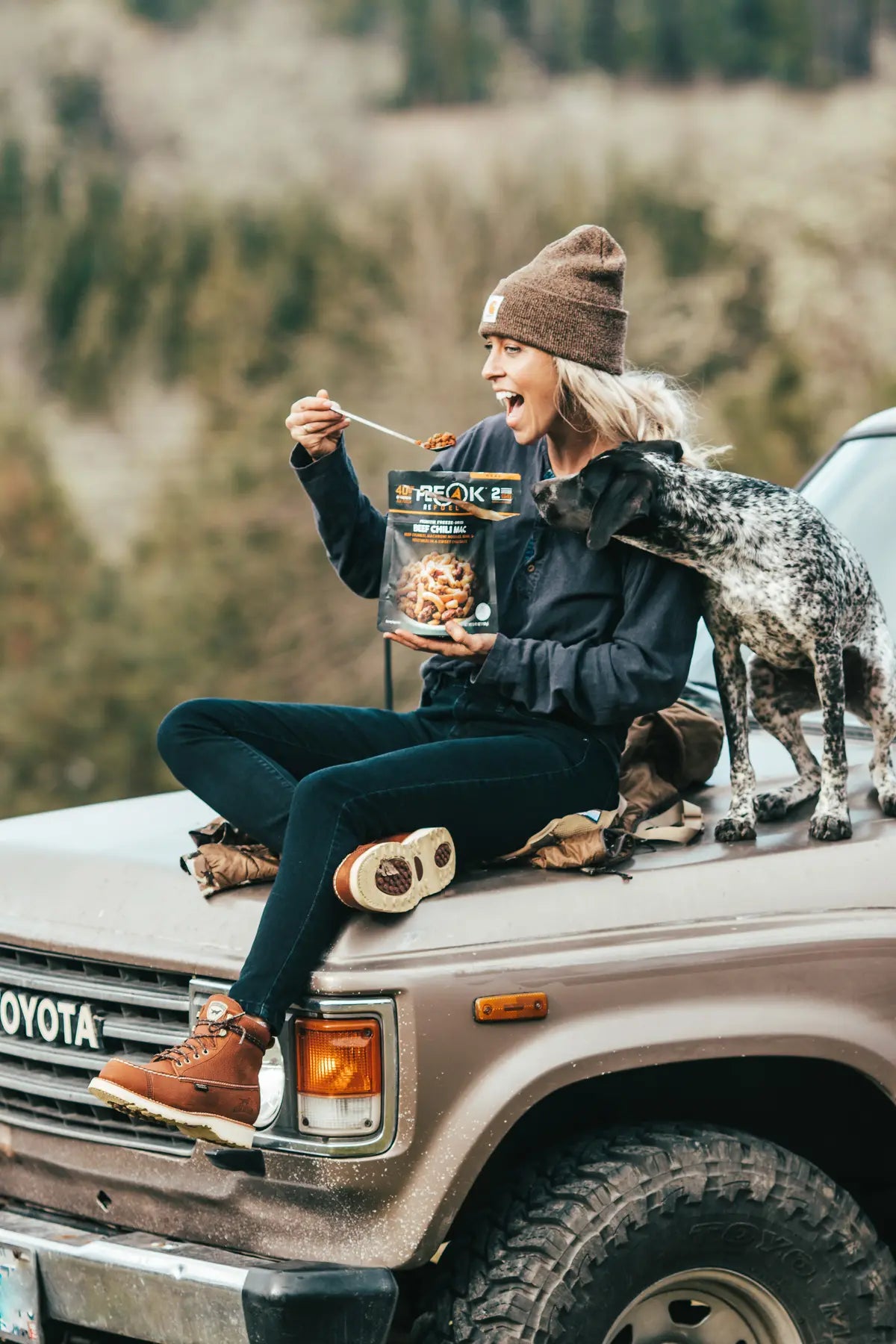 Person sitting on a Toyota vehicle with a dog, eating from a bag of Peak Refuel