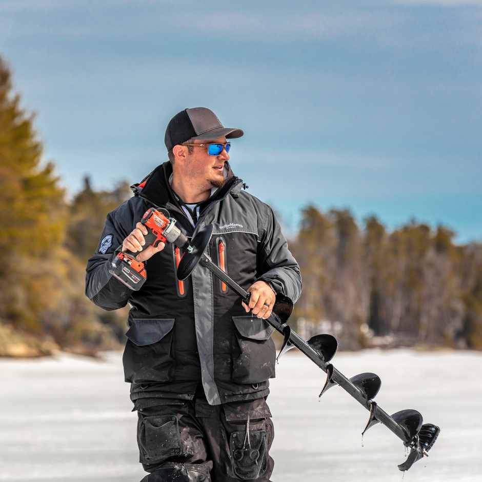 Man holding an ice auger on a frozen lake with trees in the background
