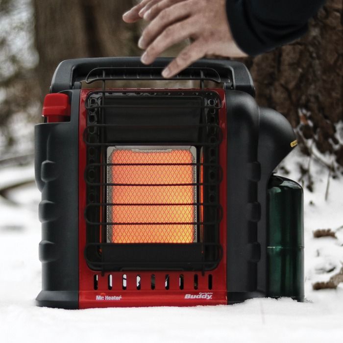 A compact, bright red portable propane heater with a fold-down handle on top and a black protective grill on the front. The heater's control knob and ignition switch are visible on the side. It sits on a flat surface with a small propane cylinder connected at the bottom.