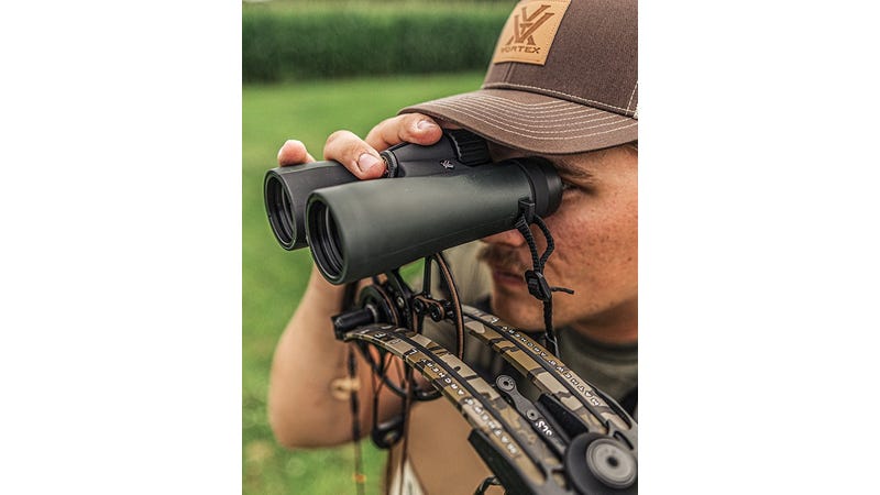 Person using binoculars with a bow and arrow setup in an outdoor setting