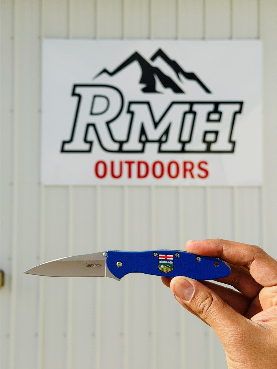 Hand holding a blue folding knife in front of an RMH Outdoors sign.