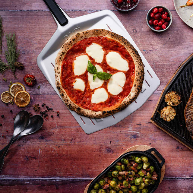 Wooden table with a pizza on a white pan, surrounded by various food items and utensils.