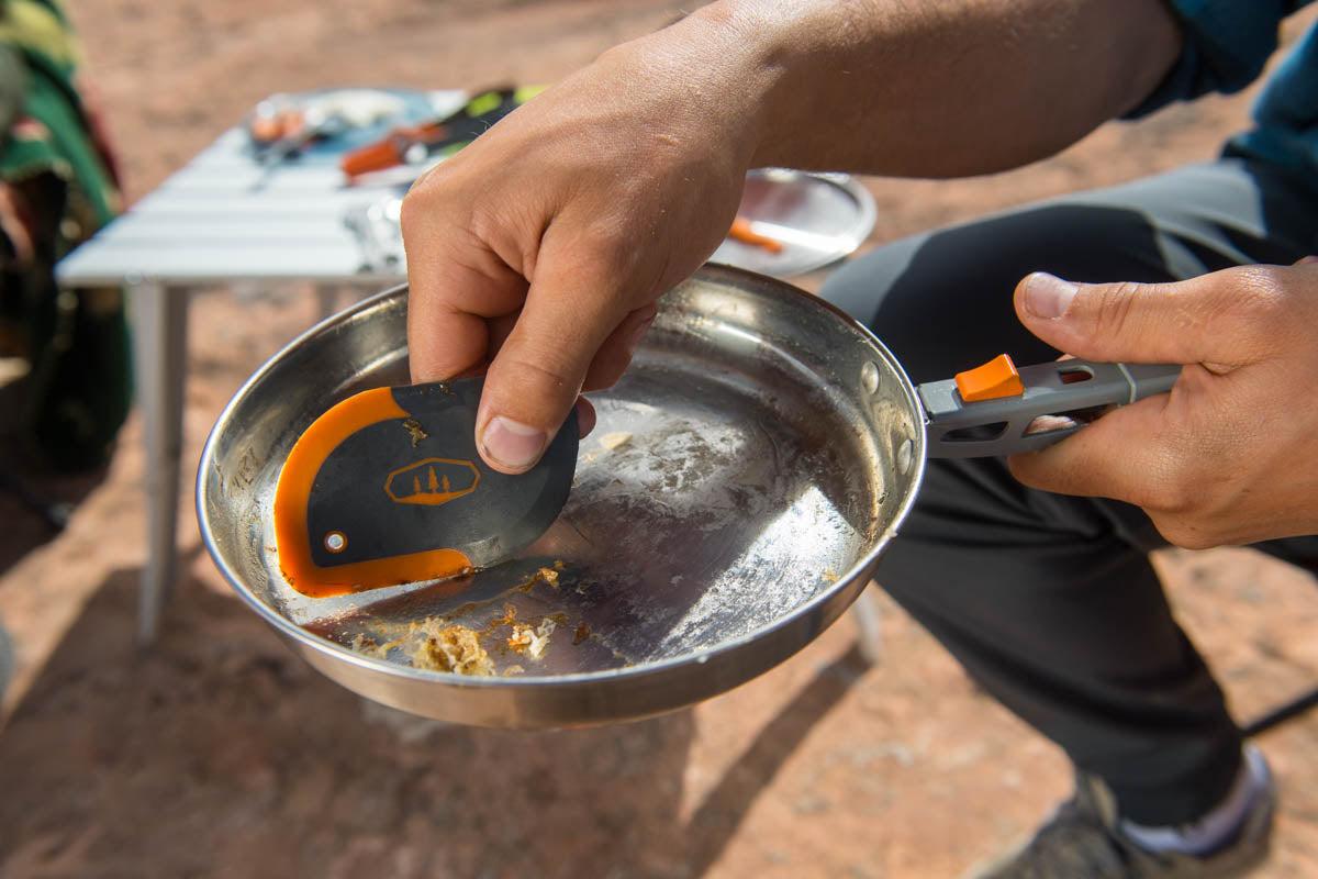 Scraper being used to clean a camping pan or pot outdoors.