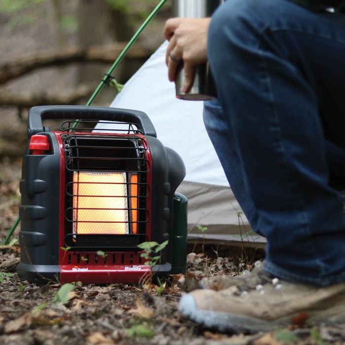 The propane heater set up in an outdoor setting, emitting radiant heat. It is placed on a camping ground with trees and tents in the background, showing its portability and outdoor functionality.