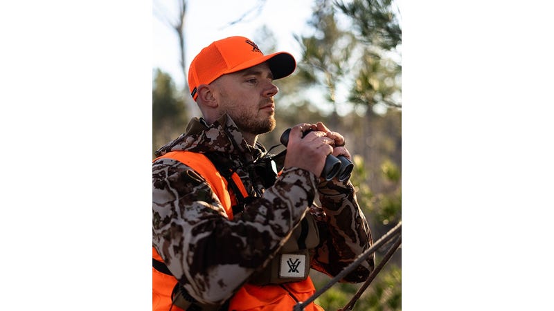 Hunter in camouflage jacket and orange vest holding a bow with trees in the background